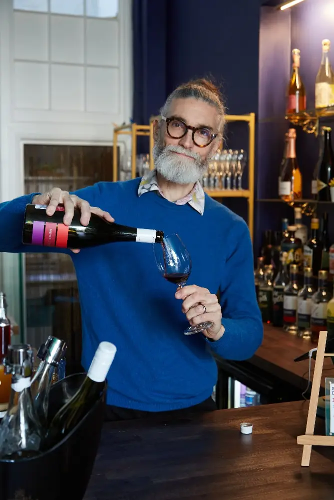 Photo of Josh Kelly - gray beard, glasses - pouring a glass of wine in a bar.