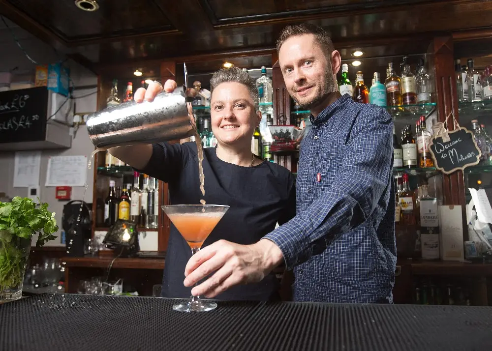 Photo of Laura and Jussi in a bar, Laura is pouring an orange cocktail from a shaker to a glass